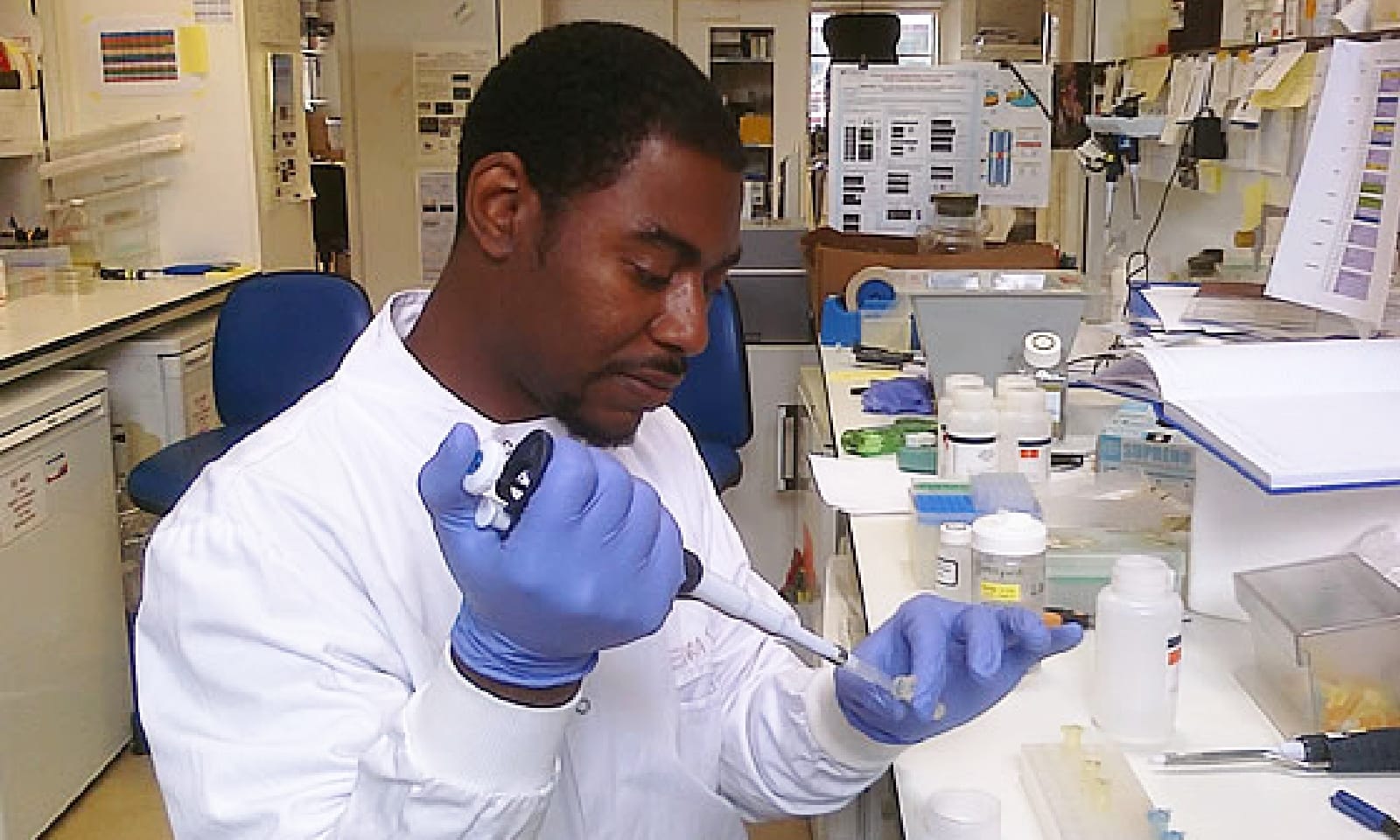 A young man in a white lab coat and gloves works with equipment in a lab.