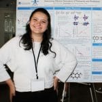 A chemistry student at Cal State Fullerton stands smiling in front of her research poster at a research symposium. She wears a white sweater and a name badge, showcasing her chemistry work.