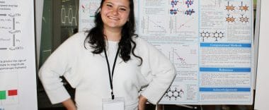 A chemistry student at Cal State Fullerton stands smiling in front of her research poster at a research symposium. She wears a white sweater and a name badge, showcasing her chemistry work.